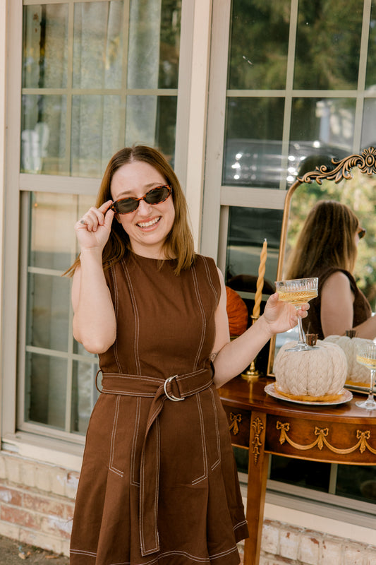 Woman in a brown dress holding sunglasses and a glass, standing in front of a window with another person in the background.