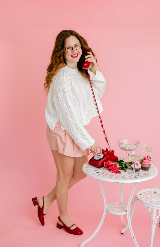 Woman in a white blouse and pink skirt holding a red vintage phone against a pink background