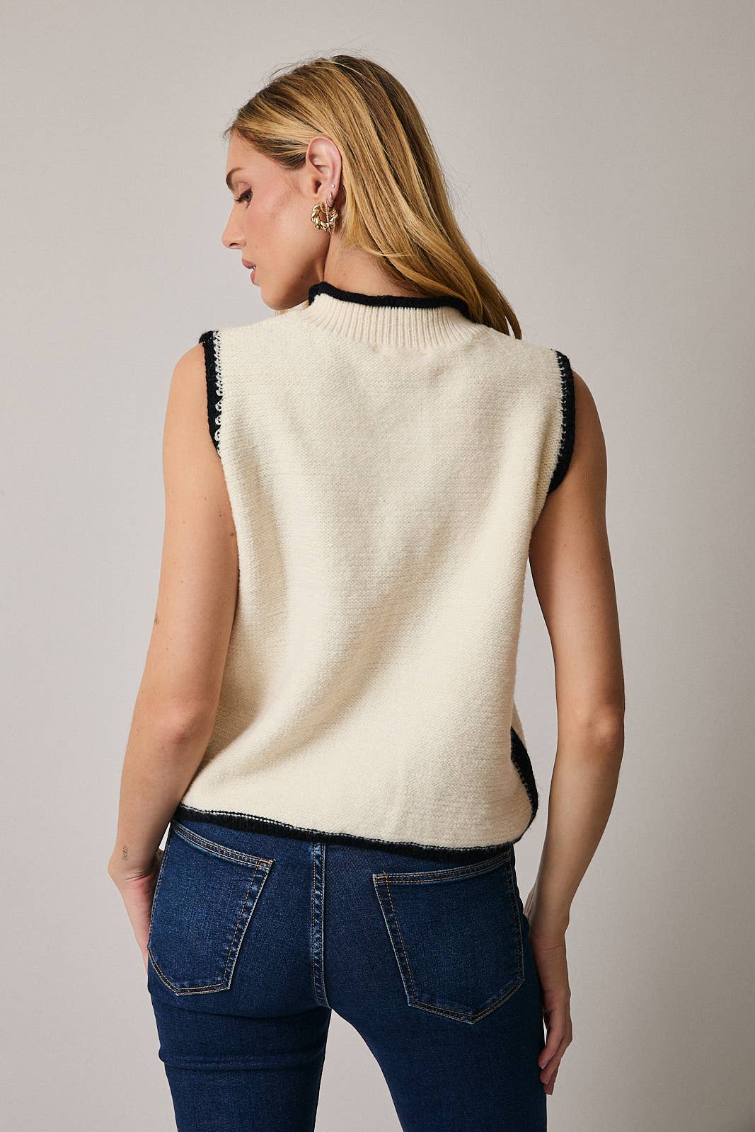Woman wearing a cream sleeveless sweater with black trim against a plain background
