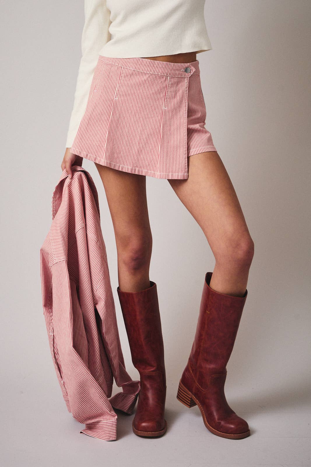 Person wearing a pink skirt and red boots holding a matching pink jacket on a plain background