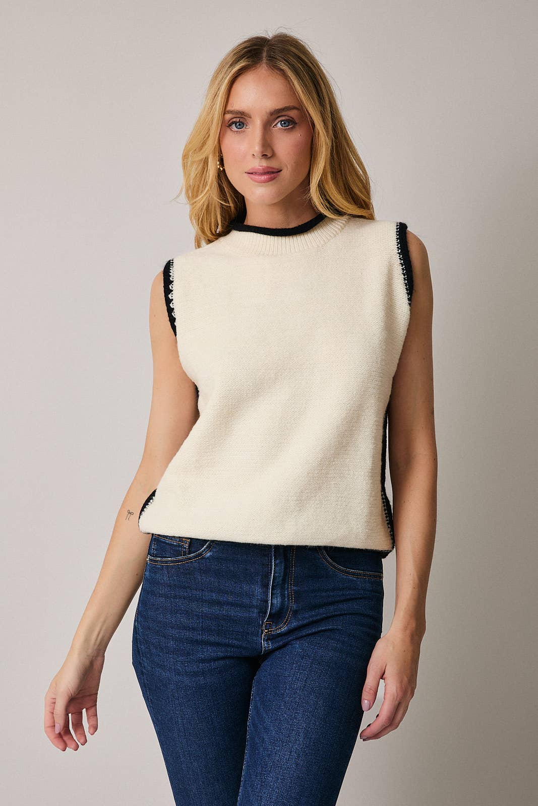 Woman wearing a cream sleeveless sweater with black trim against a neutral background
