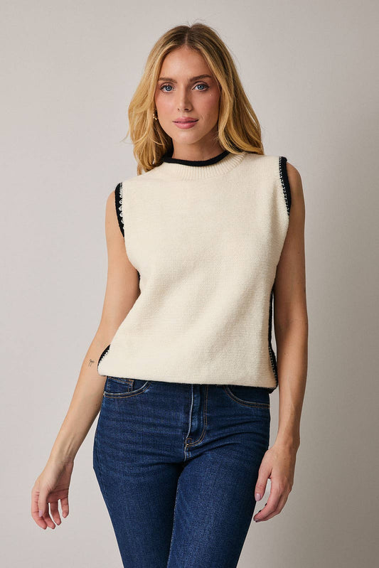 Woman wearing a cream sleeveless sweater with black trim against a neutral background