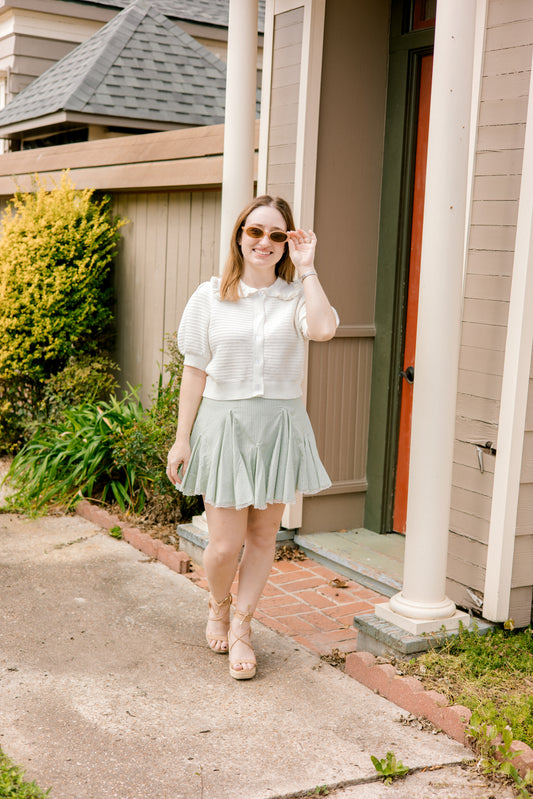 Woman wearing a white cardigan with collar and light green skirt standing outdoors.