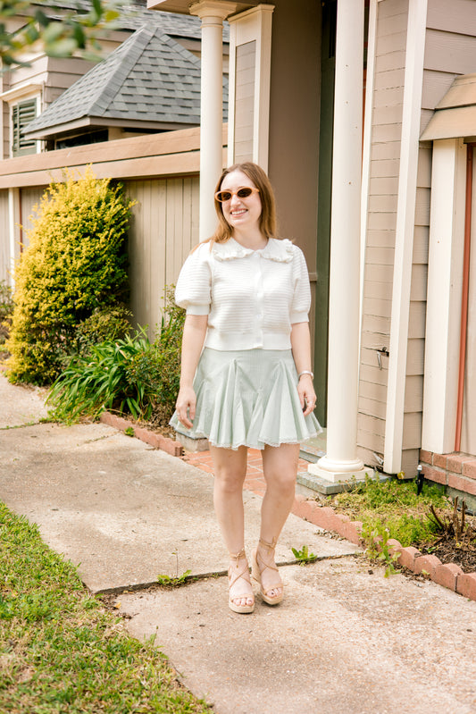 Woman wearing a white cardigan with collar and light green skirt standing outdoors.