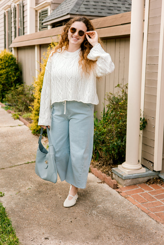 Woman in a white blouse and light stripe blue pants standing on a sidewalk.