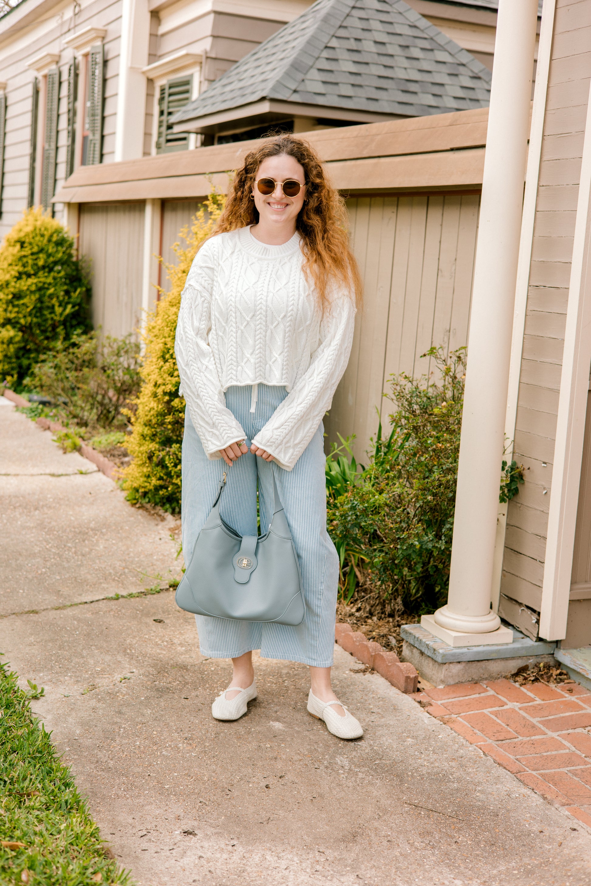 Woman standing on a sidewalk wearing a white sweater and light blue pants, holding a light blue handbag.