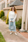 Woman standing on a sidewalk wearing a white sweater and light blue pants, holding a light blue handbag.