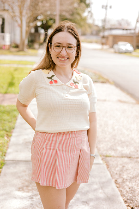 Woman wearing a white blouse with cherry designs and a pink skirt on a sidewalk.