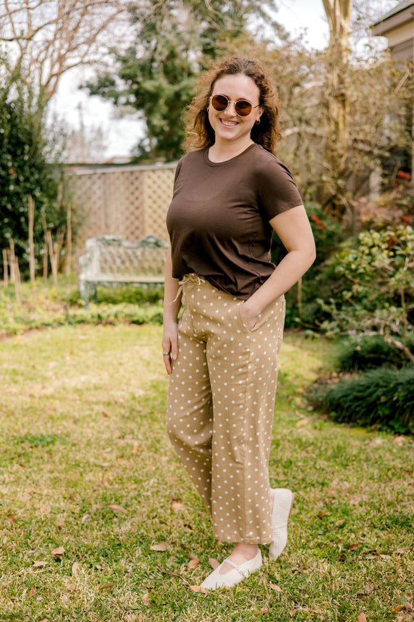 Woman standing outdoors in a garden wearing a brown top and tan/khaki polka dot pants.