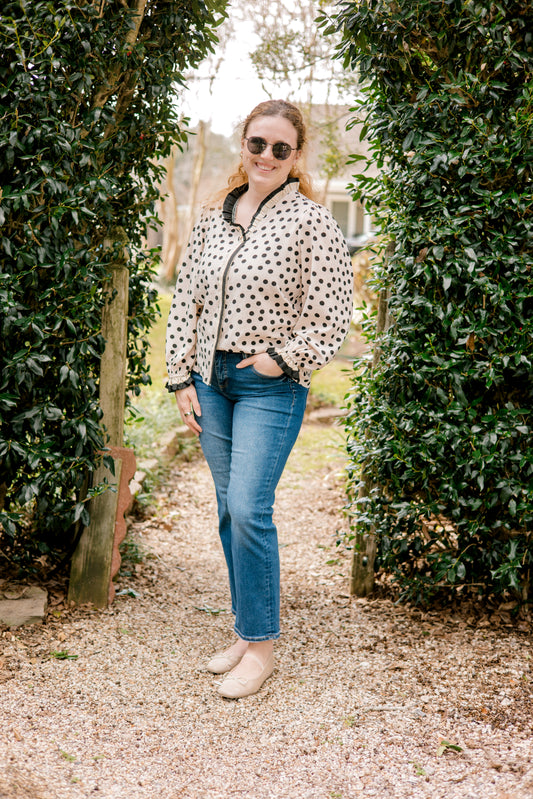 Woman in a polka dot shirt and jeans standing between two rows of green shrubs.