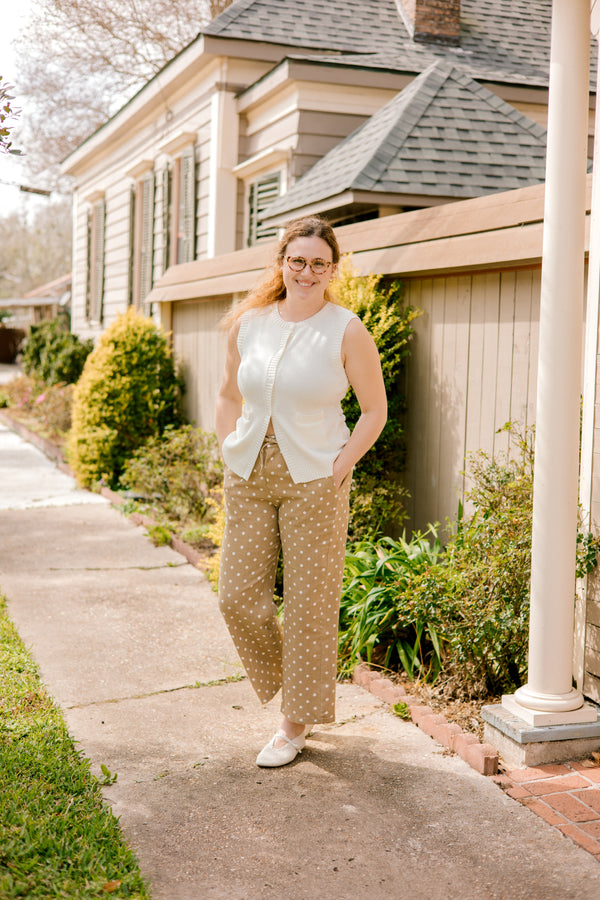 Woman standing on a sidewalk in front of a house wearing a white sleeveless top and tan khaki polka dot pants