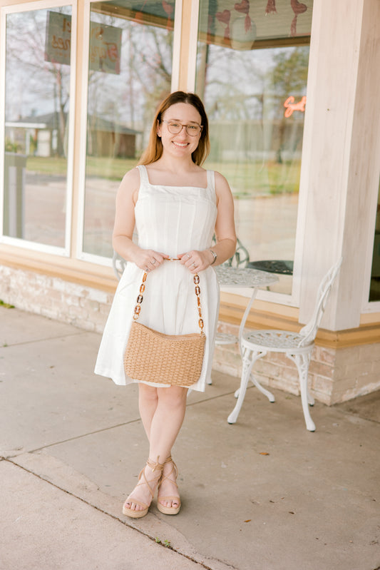Woman in a white dress with a straw bag standing outside a building.