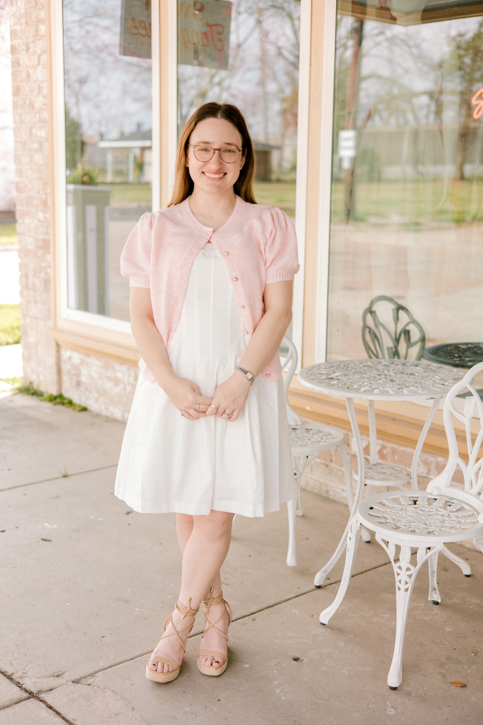 Woman in a white dress and pink cardigan standing outside a building with glass windows.