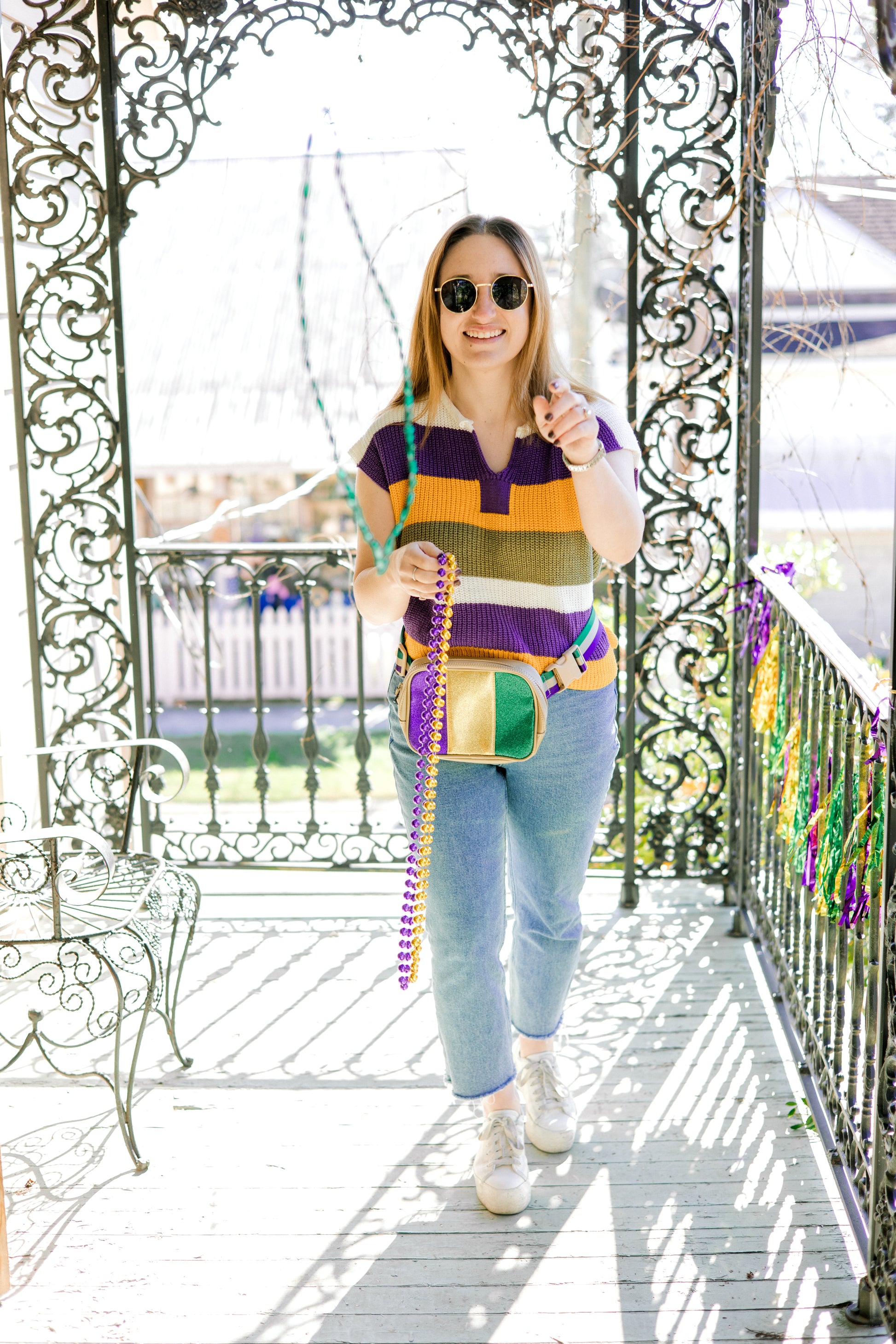 Woman on a porch with Mardi Gras decorations, wearing sunglasses and a colorful striped shirt.