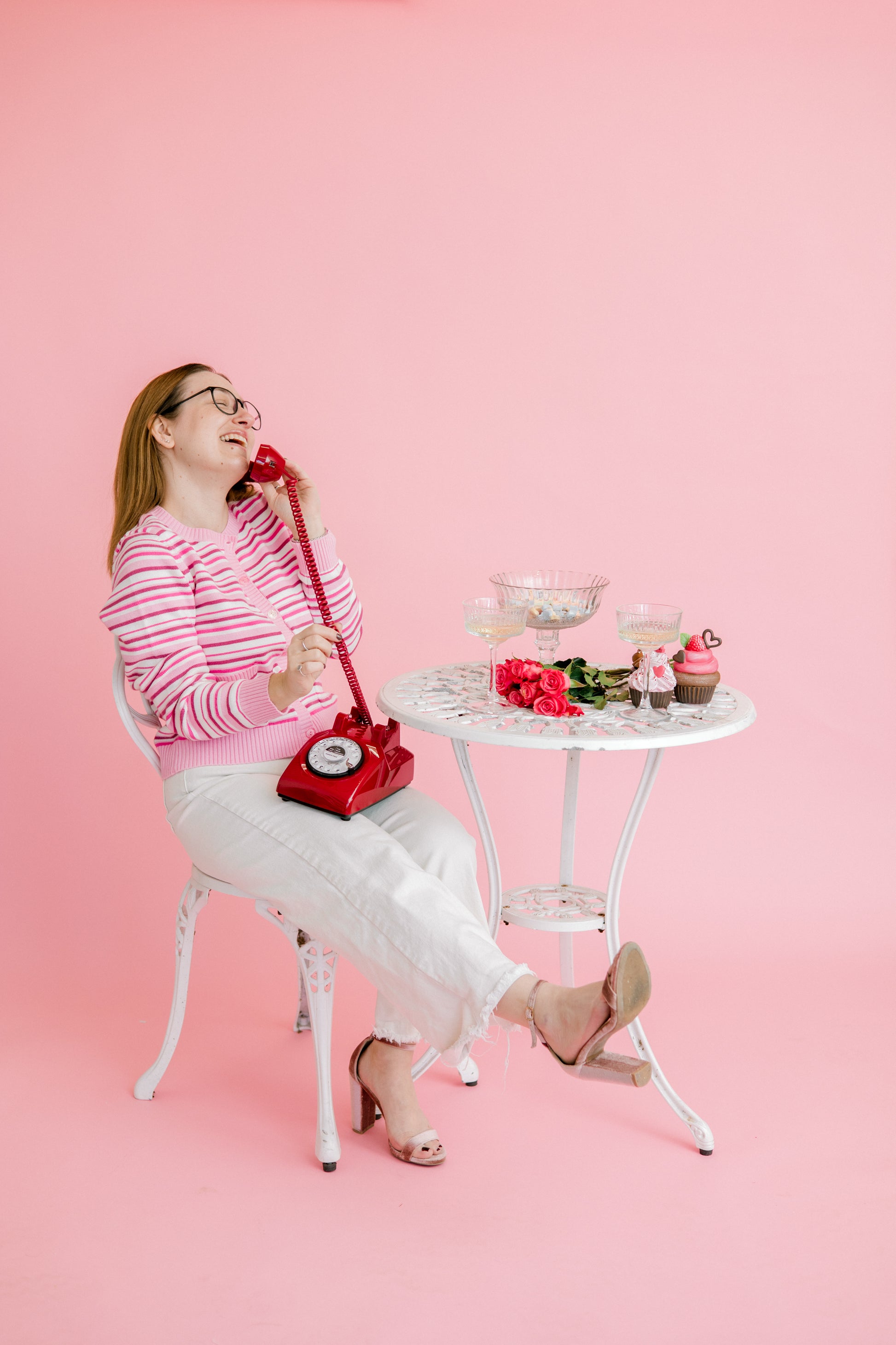 Woman sitting at a small table with a red phone and cupcakes on a pink background