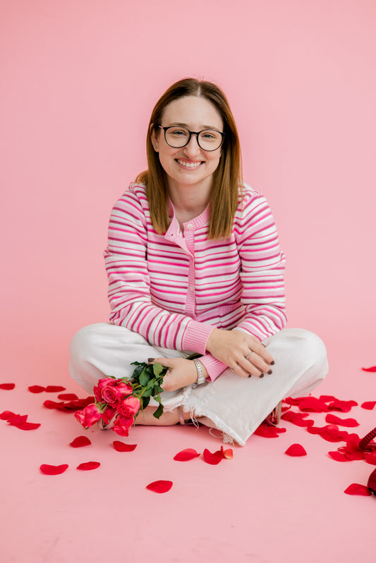 Woman sitting on a pink background with flowers and rose petals with a pink stripe cardigan on