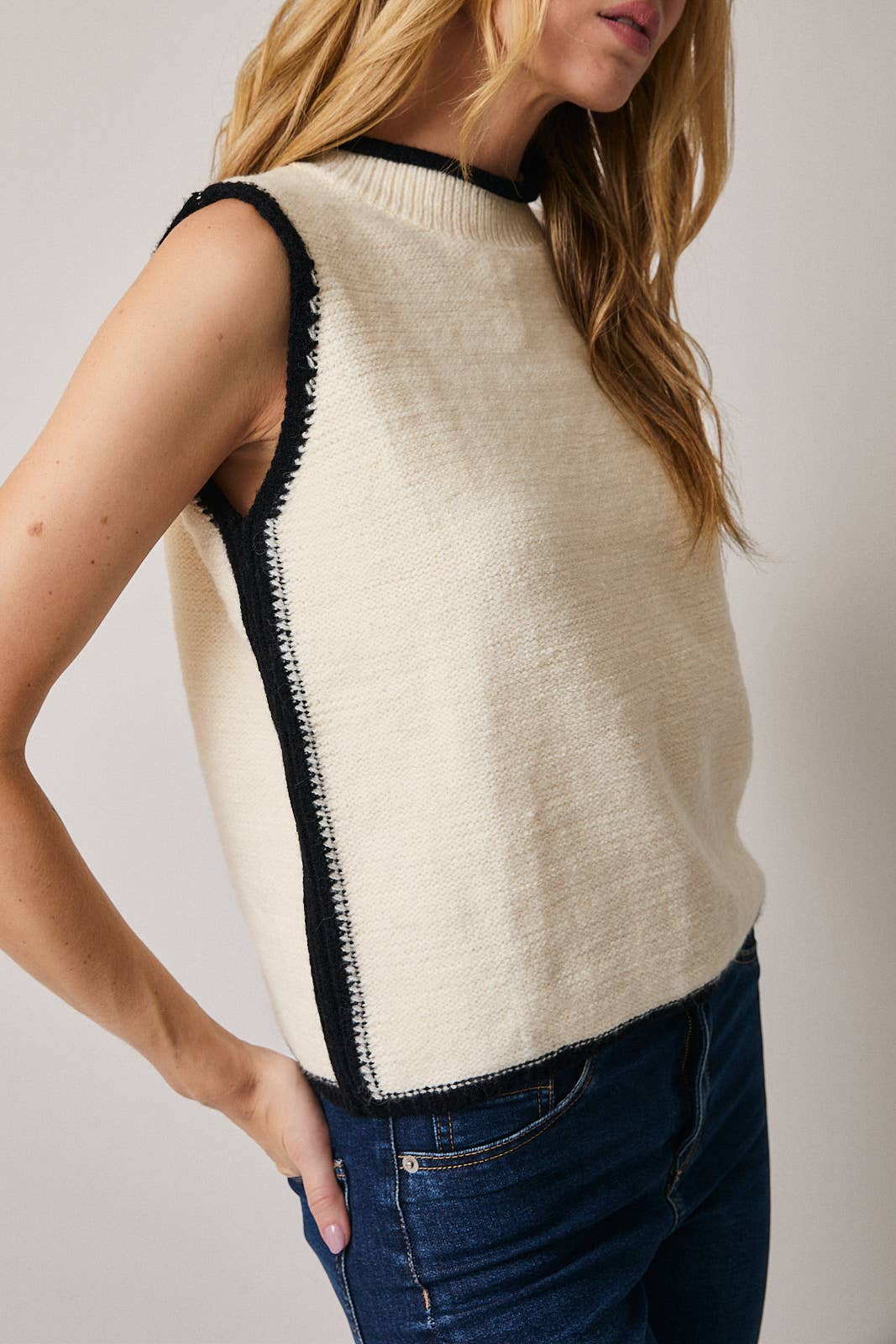 Woman wearing a cream sleeveless sweater with black trim against a plain background