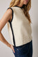 Woman wearing a cream sleeveless sweater with black trim against a plain background