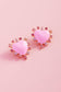 Heart-shaped pink earrings with gemstone accents on a pink background