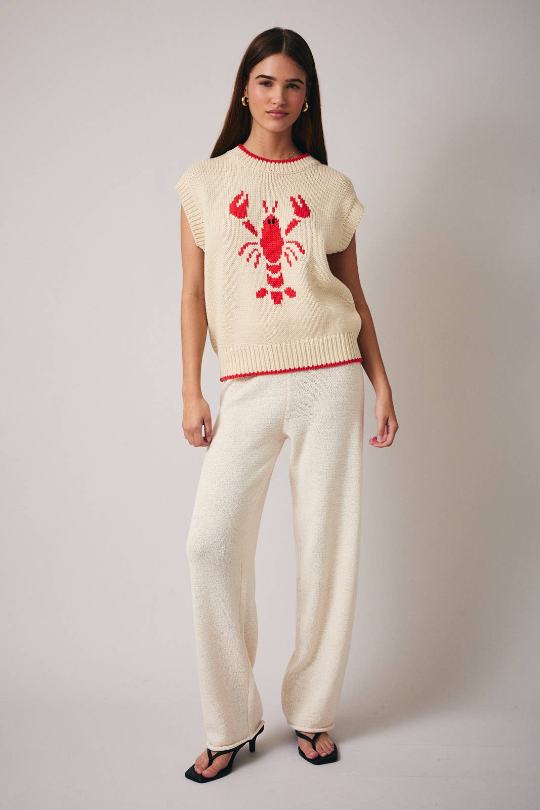 Woman wearing a cream sweater with a red lobster design and matching pants on a plain background